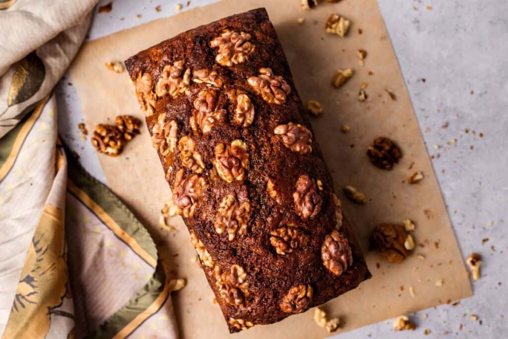 A loaf of walnut bread sits on a piece of parchment paper. The bread is topped with whole walnut halves and surrounded by a few loose walnuts. A patterned cloth is partially draped on the left side of the image. The background surface is light-colored.