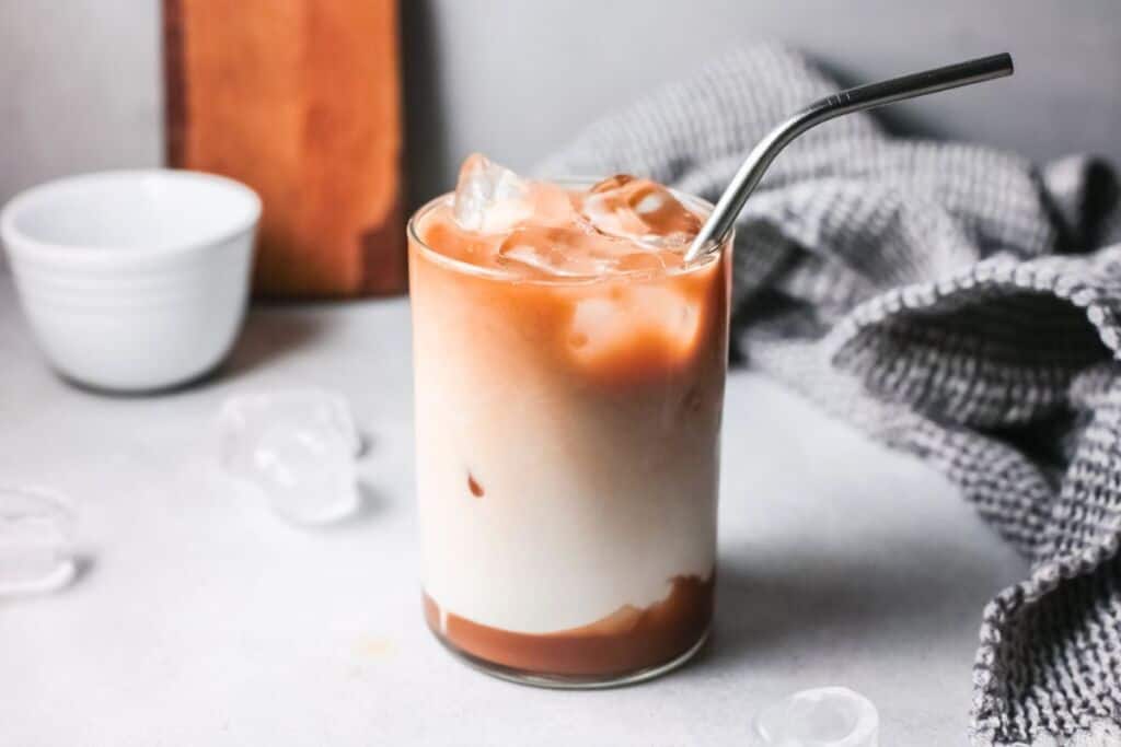 A glass of iced coffee with a metal straw sits on a light-colored surface. The drink has a visually appealing gradient from white at the bottom to light brown at the top. Ice cubes are scattered around, and a gray cloth is partially visible in the background.