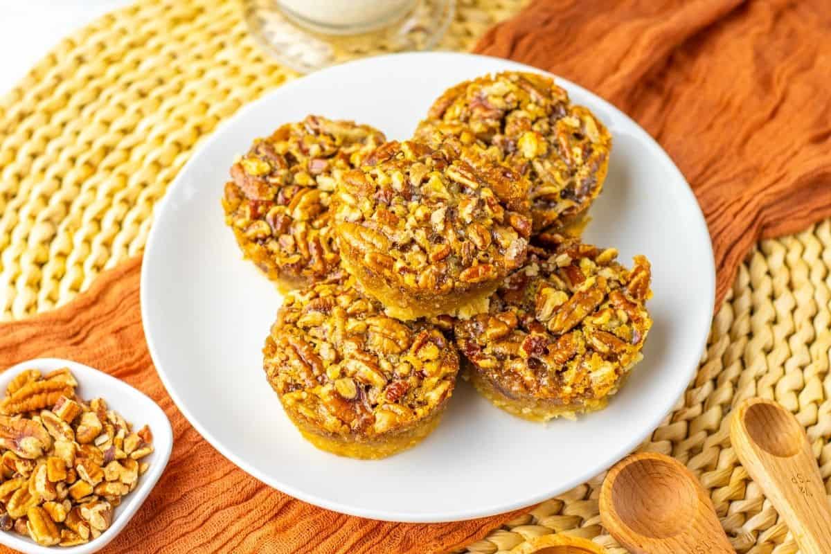 individual pecan pies on plate with bowl of chopped pecans alongside.